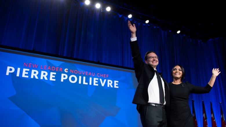 A man and a woman stand on a stage and wave.