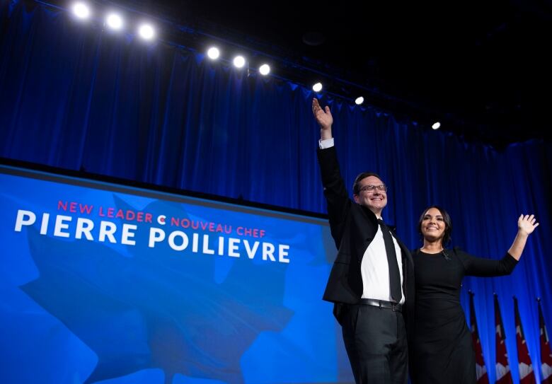 A man and a woman stand on a stage and wave.