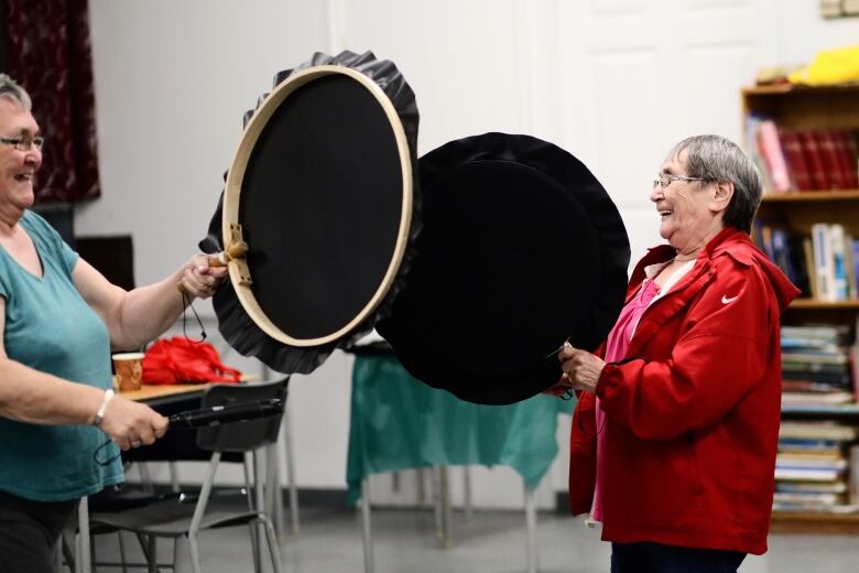 Two women holding large black drums smile and laugh at each other. 