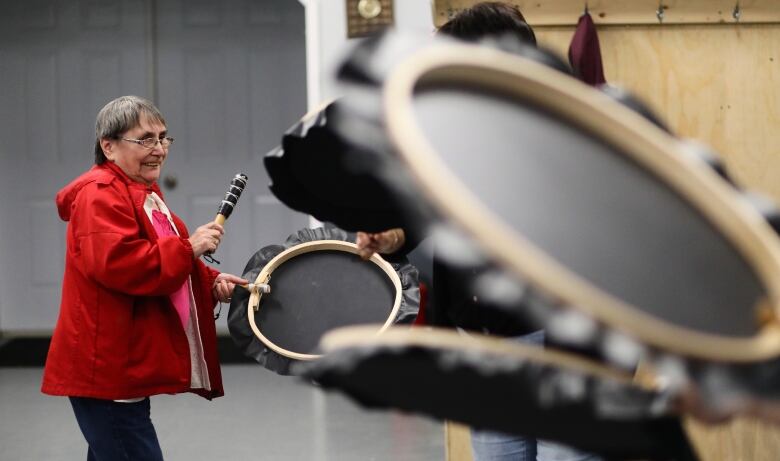 A woman in a red jacket drums while smiling and other black drums are in the frame. 