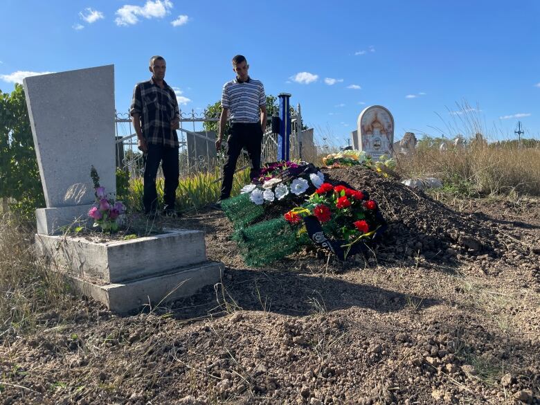 Two men stand over a gravesite covered in flowers. 