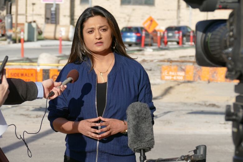 A woman stands speaking into microphones, with a construction site behind her.