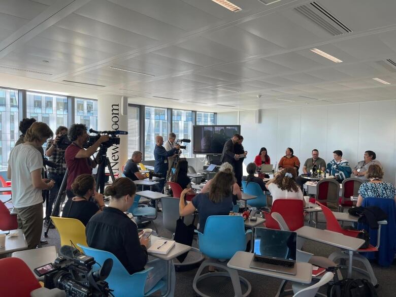 A room full of people with cameras and notepads watch a line of seated individuals.