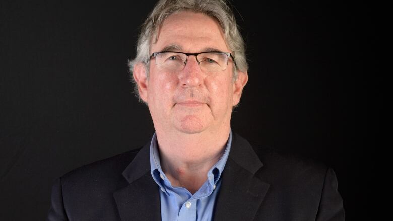 An grey-haired individual wearing a blue shirt and navy jacket. 