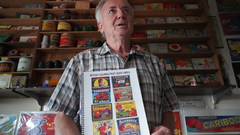 A man holds up a book with multicoloured fruit labels, with dozens of curios behind him on shelves.