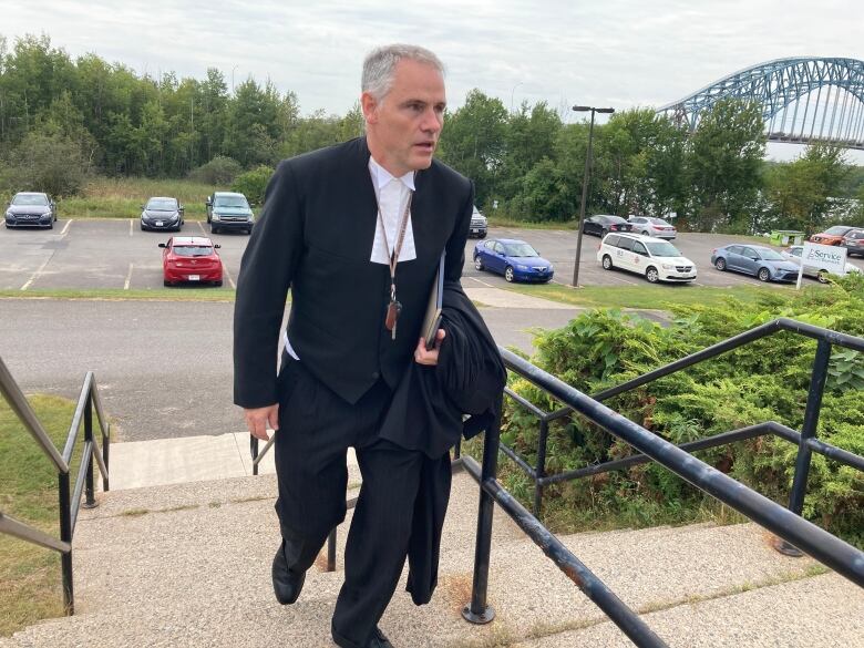 A man with short grey hair, wearing lawyer courtroom garb, climbs a concrete staircase.