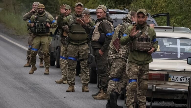 Nine men in camouflage military fatigues, with blue bands around their arms and legs, stand on the side of the road next to two parked cars. Several of them are looking toward the camera and waving. 