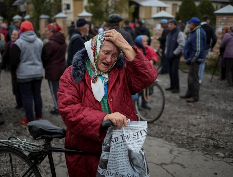 An elderly woman in a winter coat and babushka stands in the street holding up a bicycle with one hand, and placing the other hand on her head. She has a distressed expression on her face, and her mouth is agape. Many people are standing around in the background. 