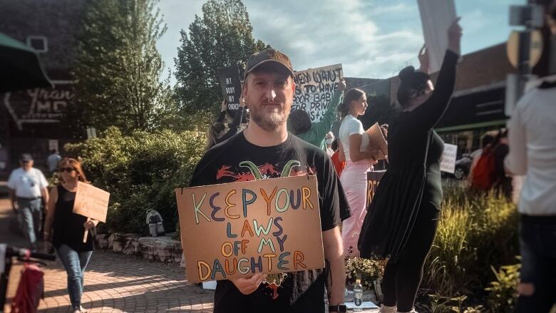 A man stands amongst other protesters in Sault Ste. Marie, Michigan holding a sign that reads 'Keep your laws off my daughter.'