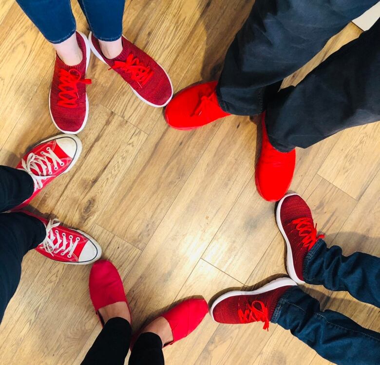 Five pairs of feet in red shoes create a star shape on a wooden floor.