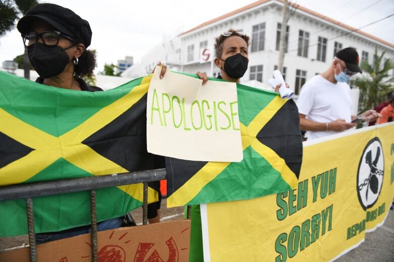 Three people holding signs in a line behind a metal guard fence with a yellow banner hanging on it.