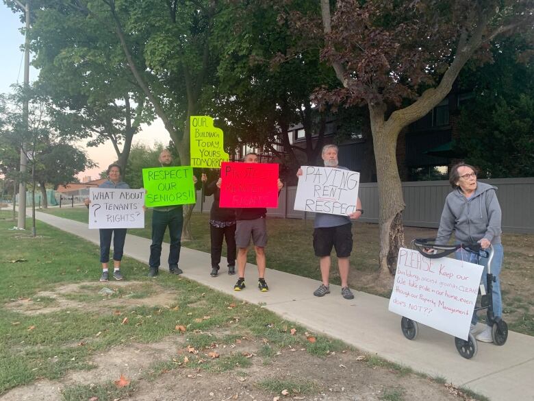 Protestors stand on a sidewalk holding signs reading 