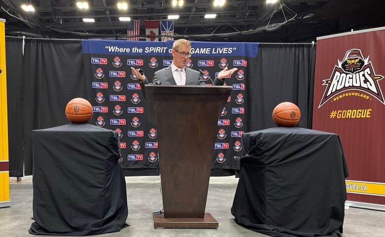 A wide shot of a person speaking at a black podium. Basketballs sit on two pedestals on his left and right. Behind him are three banners. The banner on his left and right feature logos for the Newfoundland Rogues with the hashtag #GOROGUES. Behind him is a banner featuring logos for The Basketball League.