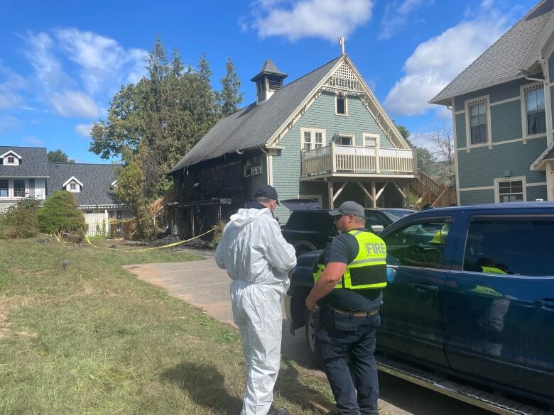 A man in protective clothing and another in a safety vest speak to each other with the burned building in the background.