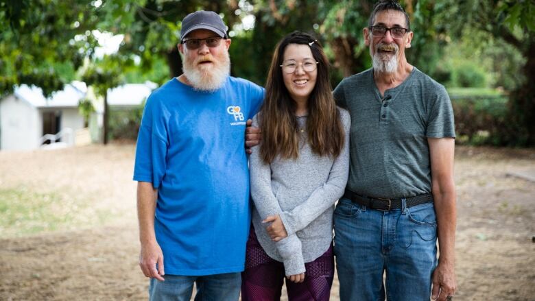 Two older white men stand in a park with their arms around a younger East Asian woman. All three are facing the camera.