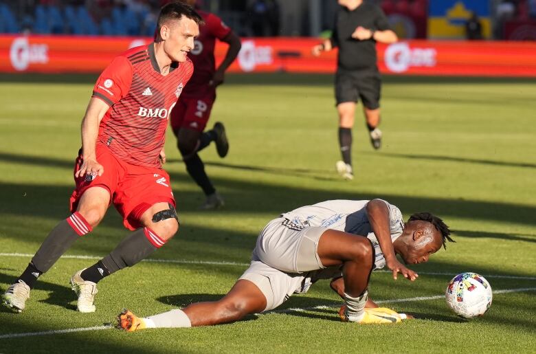 One soccer player in white is splayed on the ground near the ball, another in red stands behind him.