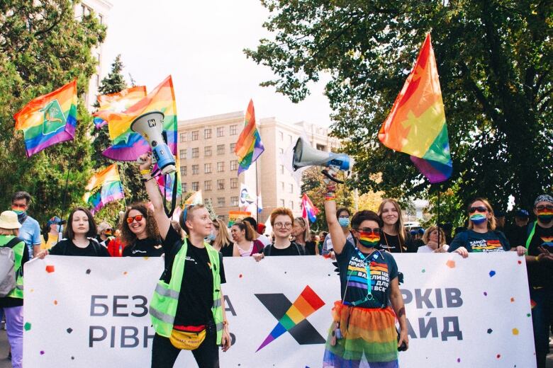 Two people stand in front of a crowd holding a white banner, while rainbow flags are raised in the air.