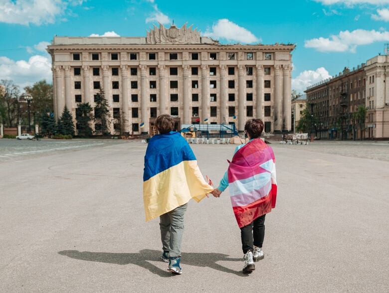 Two people with flags draped on their backs hold hands standing in front of a concrete building. 