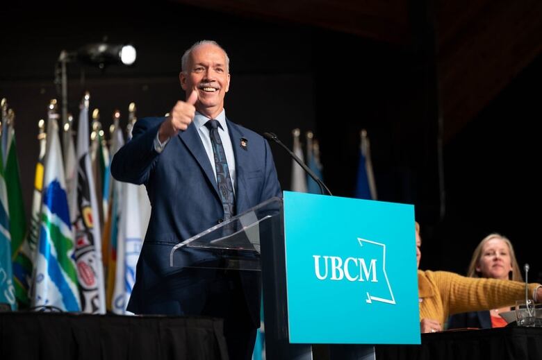 John Horgan, an old white man with a white moustache, gives a thumbs up as he stands at a podium.