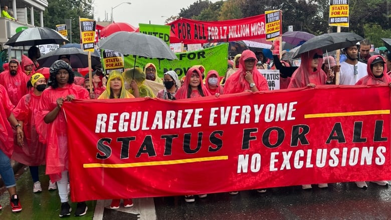 Many people wearing raincoats and holding umbrellas stand behind a red banner.