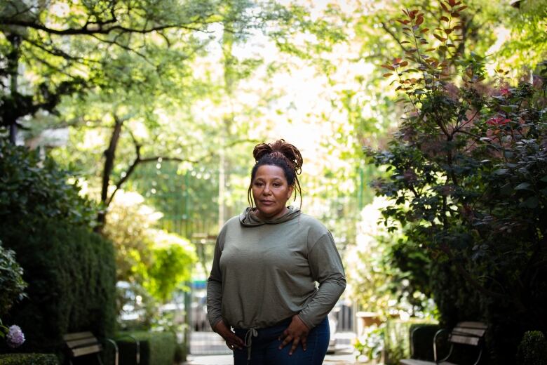 A Black woman stands among trees and greenery.