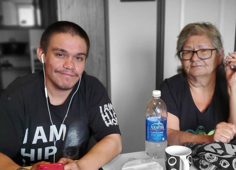 A man wearing white headphones, left, sits at a table and smiles softly. A woman wearing black glasses sits beside him at the table. 