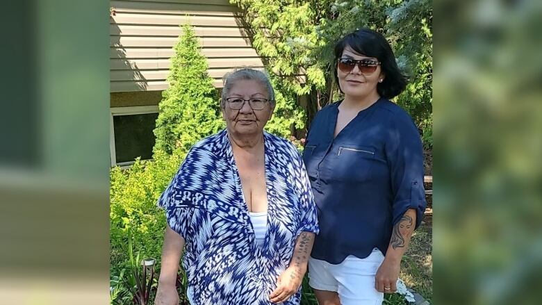 Two women stand outside of a beige home in front of green trees and bushes. 