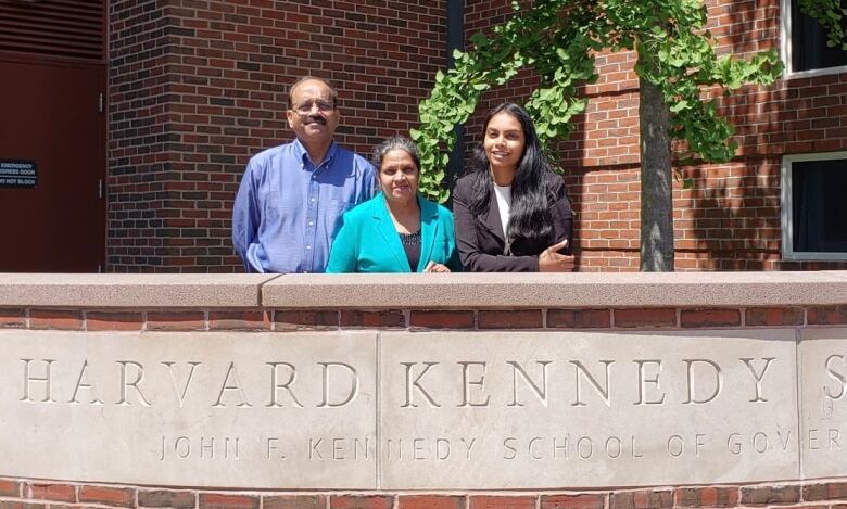 A man and two women lean on a wall above a sign for Harvard Kennedy School.