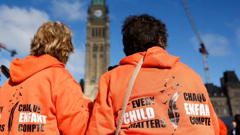 Two people seen from behind, wearing orange shirts and looking at the Peace Tower on Parliament Hill. 