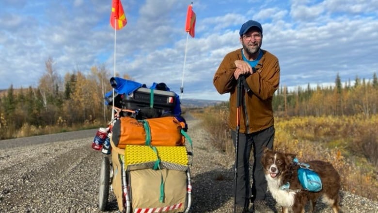 Smiling man and a small Australian Sheppard standing beside a cart on the side of the highway.  The cart has ten days worth of food and water in it, two sleeping bags, a tent and clothing.  