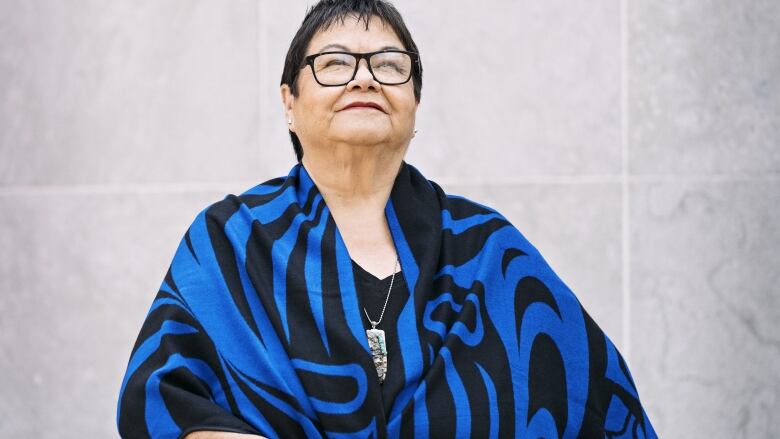 Short-haired individual wearing glasses and a blue and black shawl, looking upwards. 