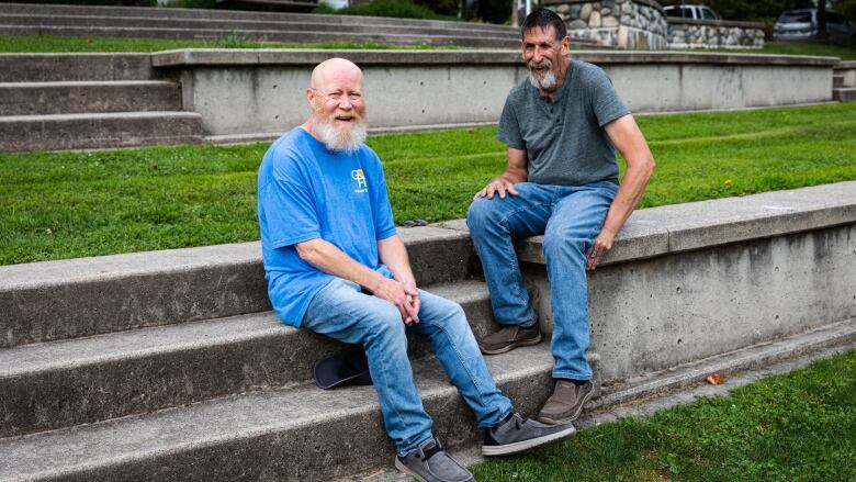 Two white men in their early 60s, both with beards and wearing jeans and t-shirts, sit on concrete steps in a park.