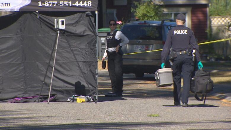 A cop walks past a black forensic tent, carrying equipment, as another cop looks on.