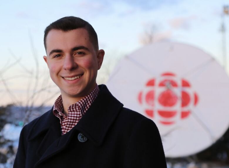 Young man short black hair wearing checkered red and white shirt and black coat standing in front of a CBC satellite dish