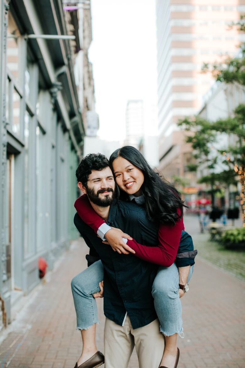 A man in a navy button up shirt with white polka dots is seen giving a woman with long, dark hair and a dark red sweater a piggy-back ride.