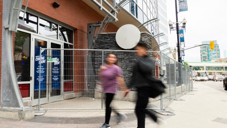 Two people walk down the sidewalk past a fence blocking off a closed downtown business.