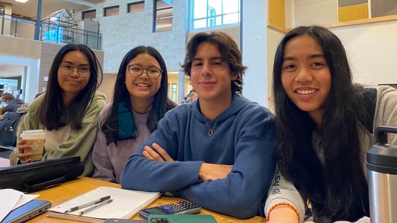 Four high school students (three teenaged girls and a teenaged boy) sit close together at a desk covered with notebooks, pens and calculators.