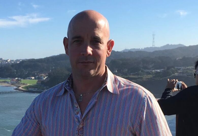 A man in a striped shirt stands in a bridge with water and hills in the background. 