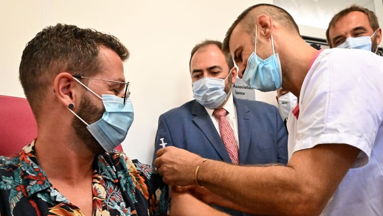 Person wearing tropical short-sleeved shirt and medical face mask sits as a health professional wearing a mask holds a vaccine needle above his arm, while others watch.