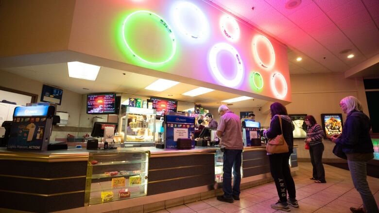 Several people line up at a concession stand covered in the rainbow-coloured glow from circular lights above.