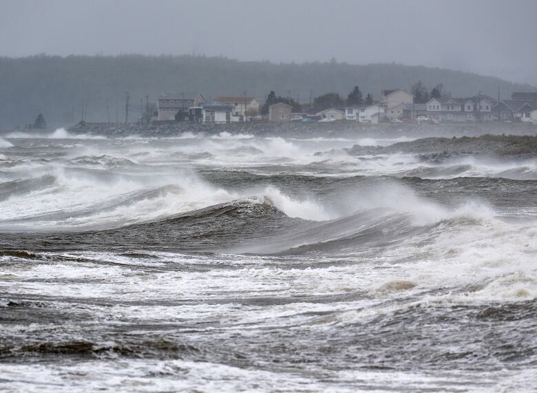 Waves and sea spray can be seen with a line of houses in the distance.