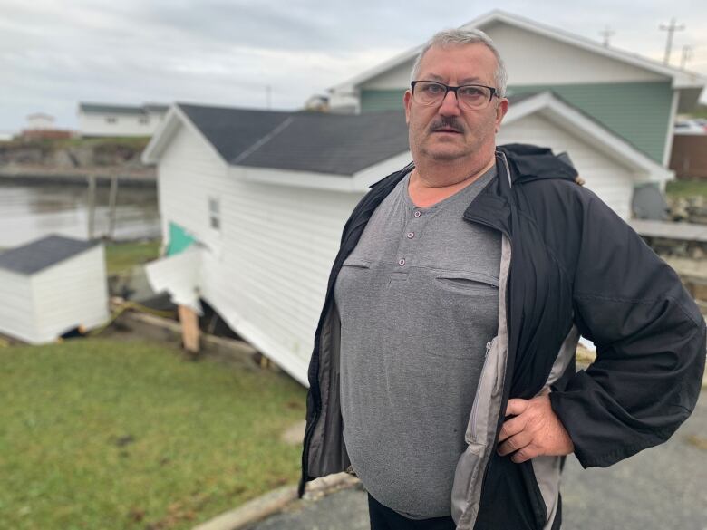 A waist-up shot of a person with his hand on his hip. Behind him, a wide house can be seen off its foundation. To the left of the house, the water is calm.