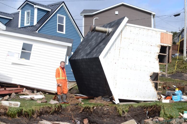 A man in coveralls stands in front of two buildings, both knocked off their foundation.