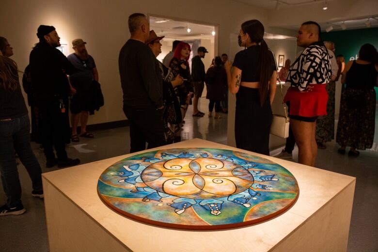 A large circular-shaped mural rests on a plinth in an art gallery, with people mingling in the background.