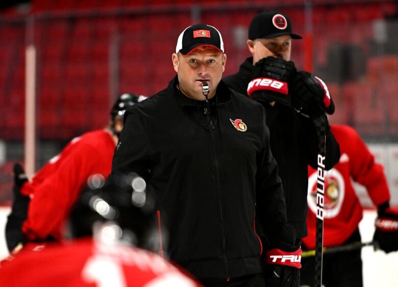 A coach in a hat and track suit with a whistle in his mouth.