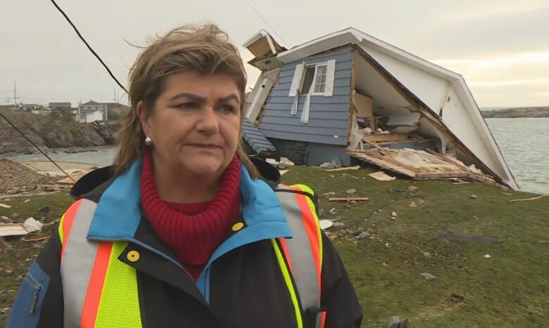 A chest-up shot of a person wearing a yellow and orange safety vest over a black and blue jacket and red turtle neck. A collapsed house can be seen behind her to the right. Behind the house is the ocean and and an overcast sky.