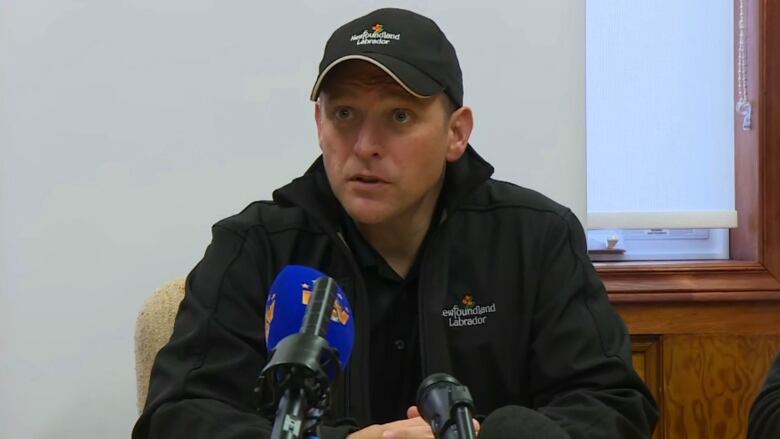 A chest-up shot of a person sitting at a table, wearing a a jacket and baseball hat with the Newfoundland and Labrador logo, speaking into a microphone.