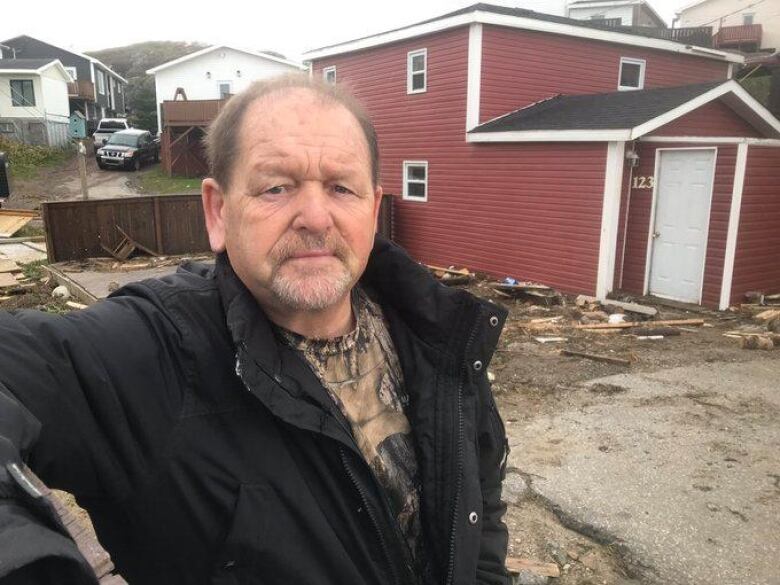 A chest-up shot of a person wearing a black jacket. Behind him to the left is a red house, with debris scattered outside.