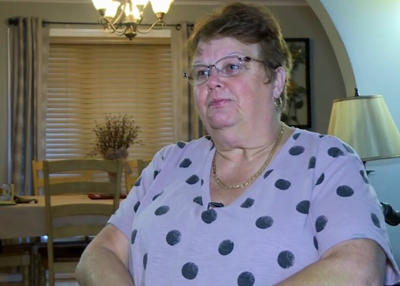 A chest-up short of a person wearing a purple polka dot shirt and glasses, sitting down. Behind the person is a lamp on the right and a dining room and window on the left.