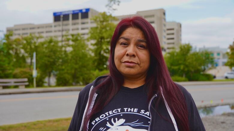 A woman with red hair look into the camera. A hospital is in the background.